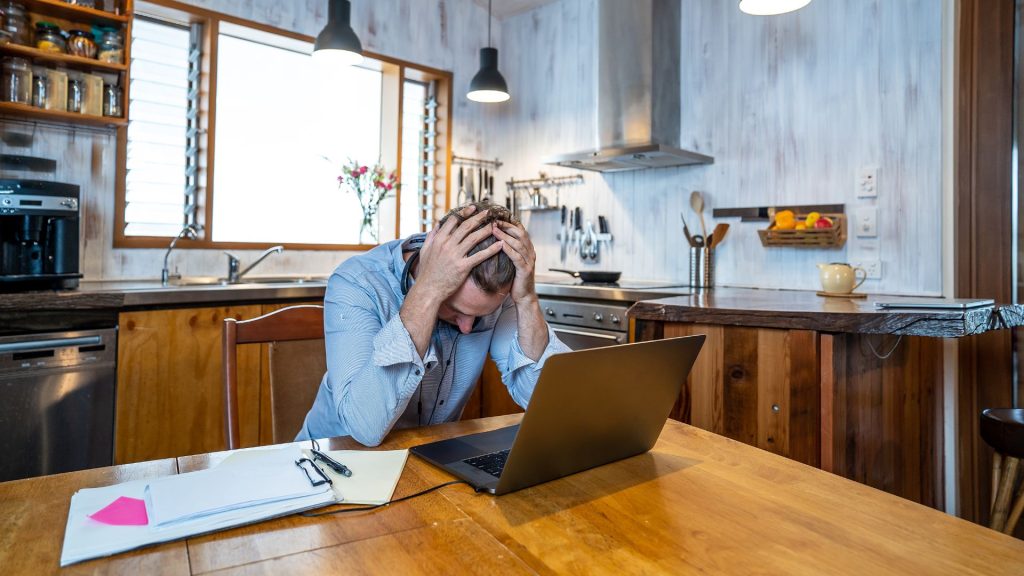 A stressed man puts his head down while avoiding looking at his laptop