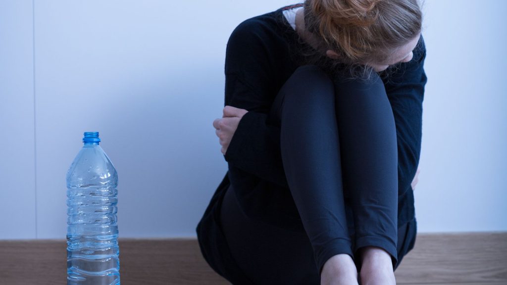 A woman holds her knees to her chest in a room with just a water bottle