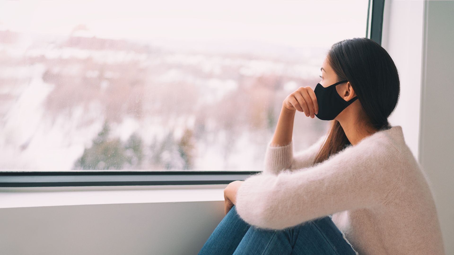 A woman with a face mask looks out through a foggy window into the distance
