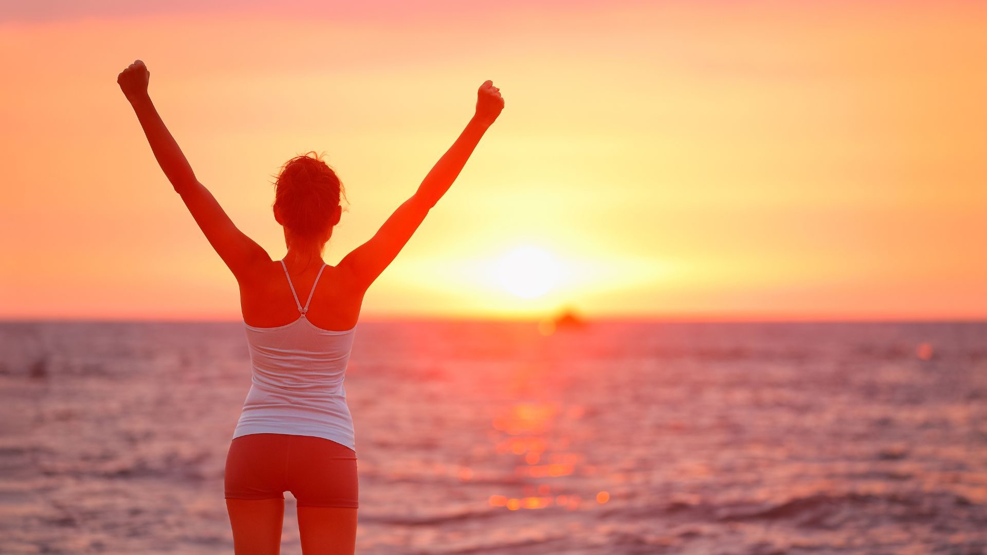 A woman stands with outstretched arms while looking at the sunset over the Pacific Ocean