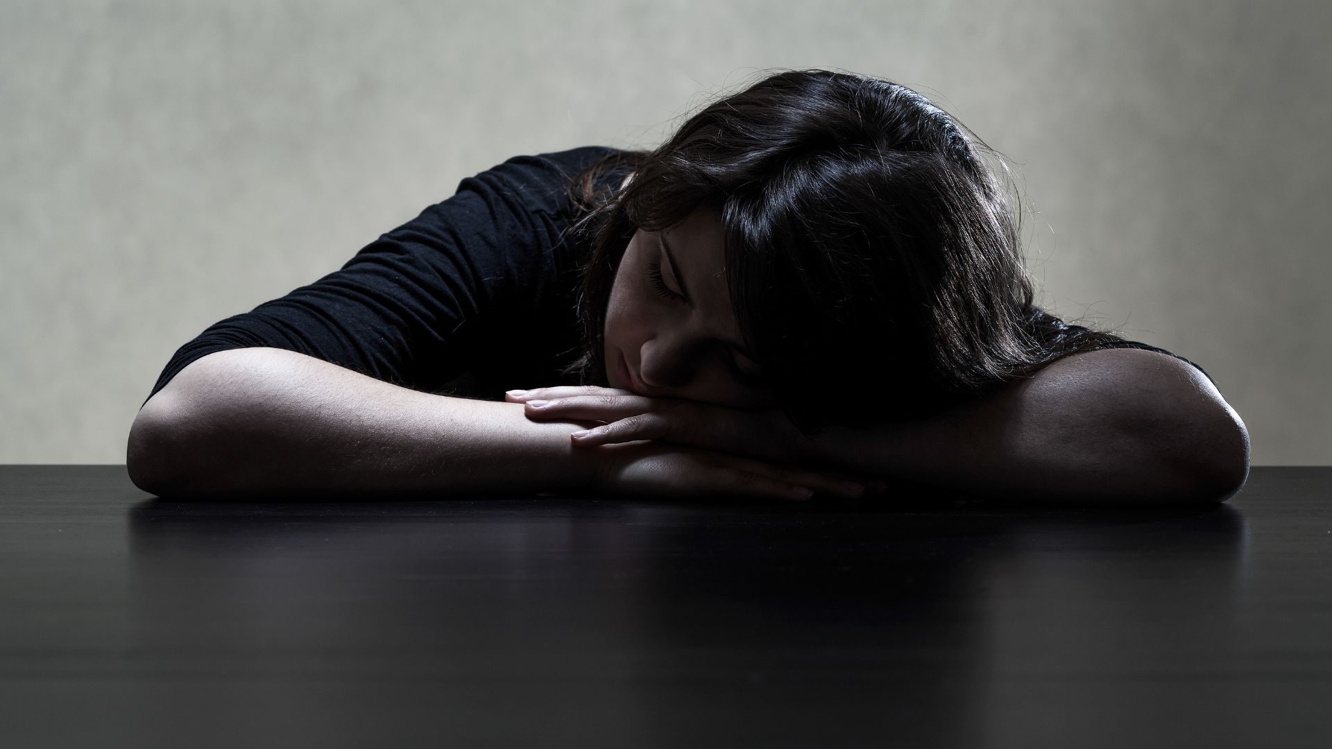 A darkly lit photograph of a young person with their head on their hands on a table