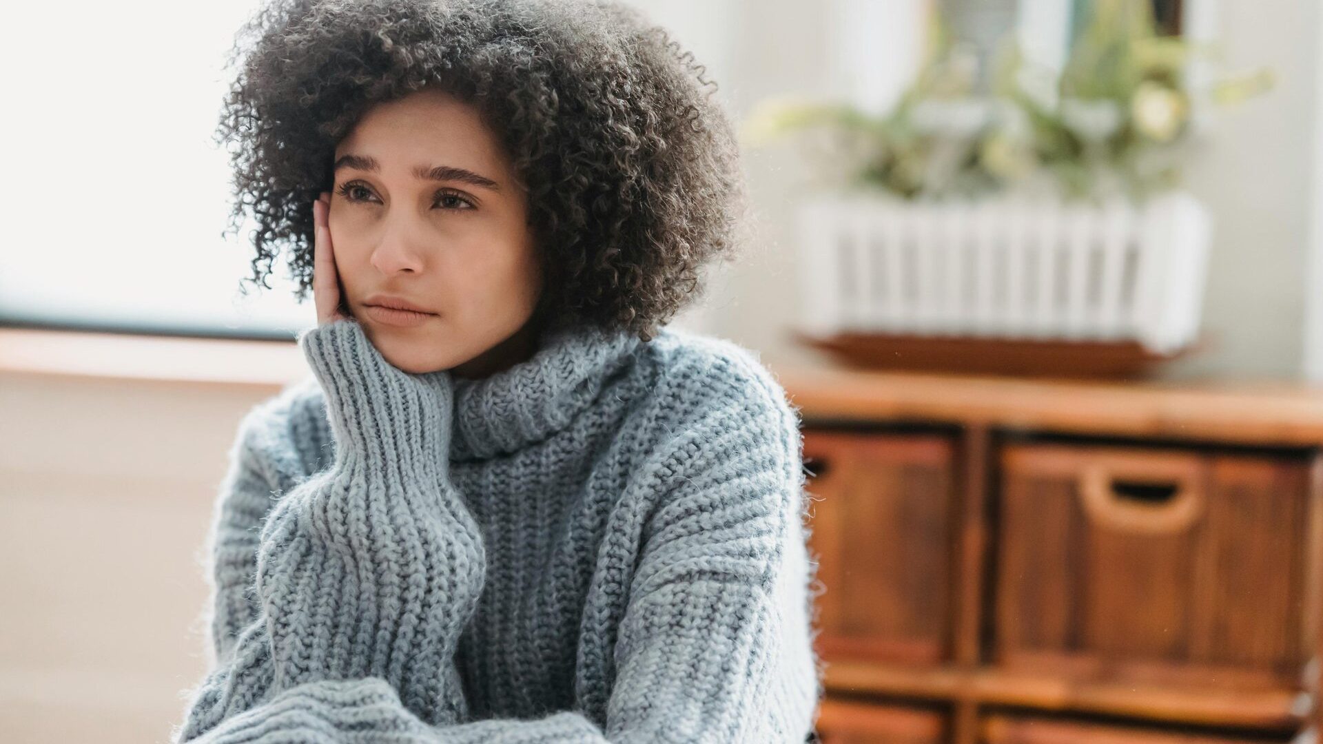 Discontent African American female leaning on hand and looking away while sitting in light room near green plant on blurred background