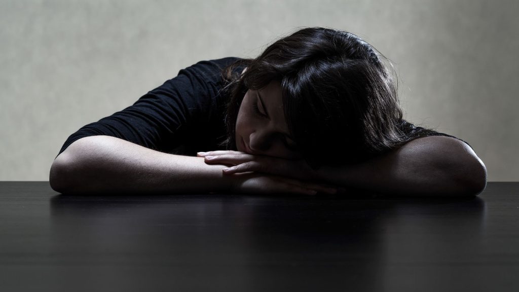 A darkly lit photograph of a young person with their head on their hands on a table