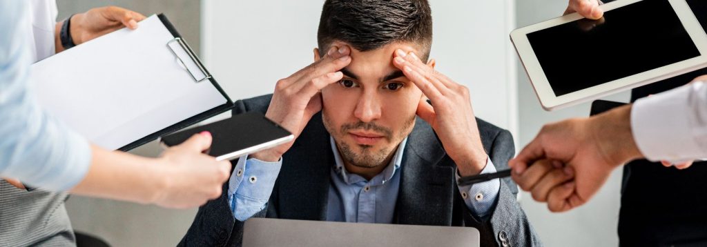 A stressed looking man surrounded by coworkers holding tablets, cell phones and paperwork