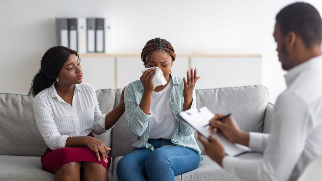 A crying woman, comforted by her daughter, in a room with a therapist