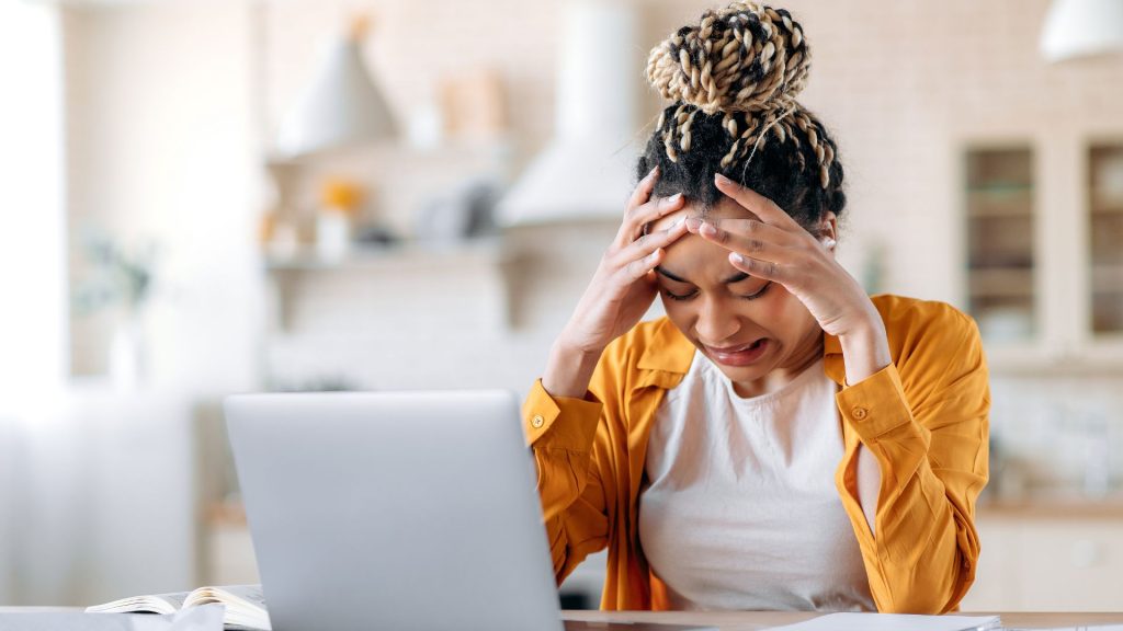 A stressed looking woman grits her teeth while looking down at the table near her open laptop